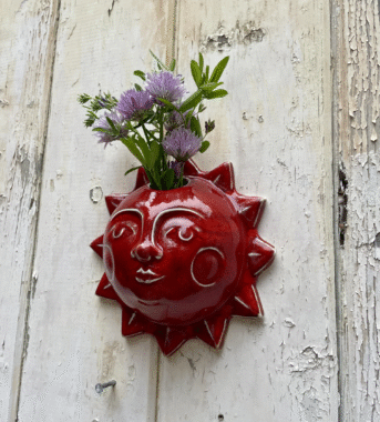 Red ceramic sun-shaped wall planter with face design holding purple flowers on weathered white wood