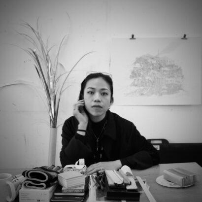 A woman looks at the camera, sat on a table laid with various objects. Behind her is an artwork on the wall.