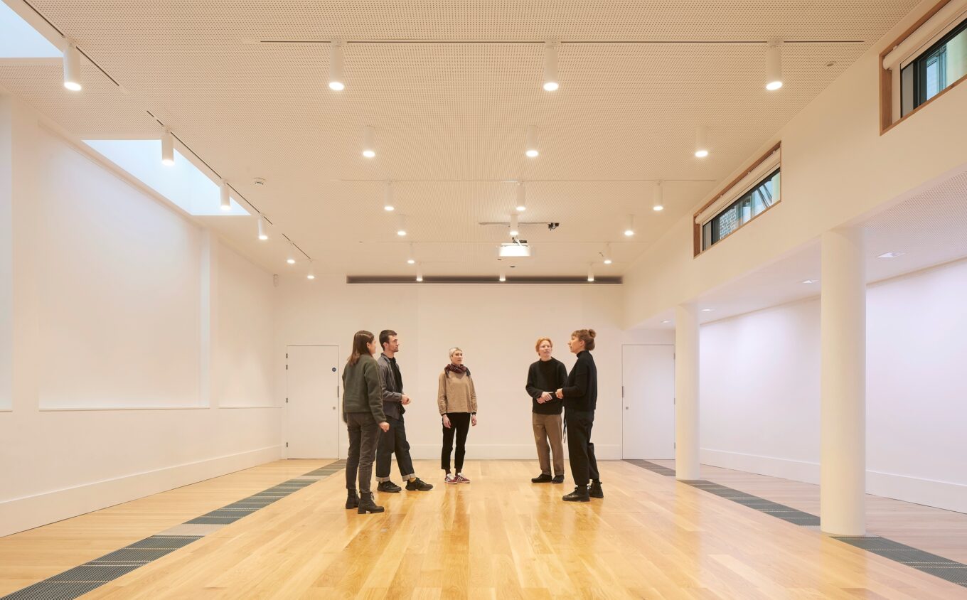 Five people standing in a brightly lit studio wall with wooden floors and white walls.