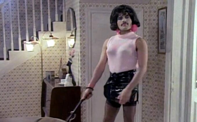 A man with a moustache, an exaggerated black bob and a tank top beneath which are fake breasts stands in a living rom, holding the handle of an upright vacuum cleaner.