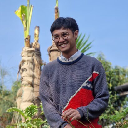 A person in a grey and red jumper stands among greenery.