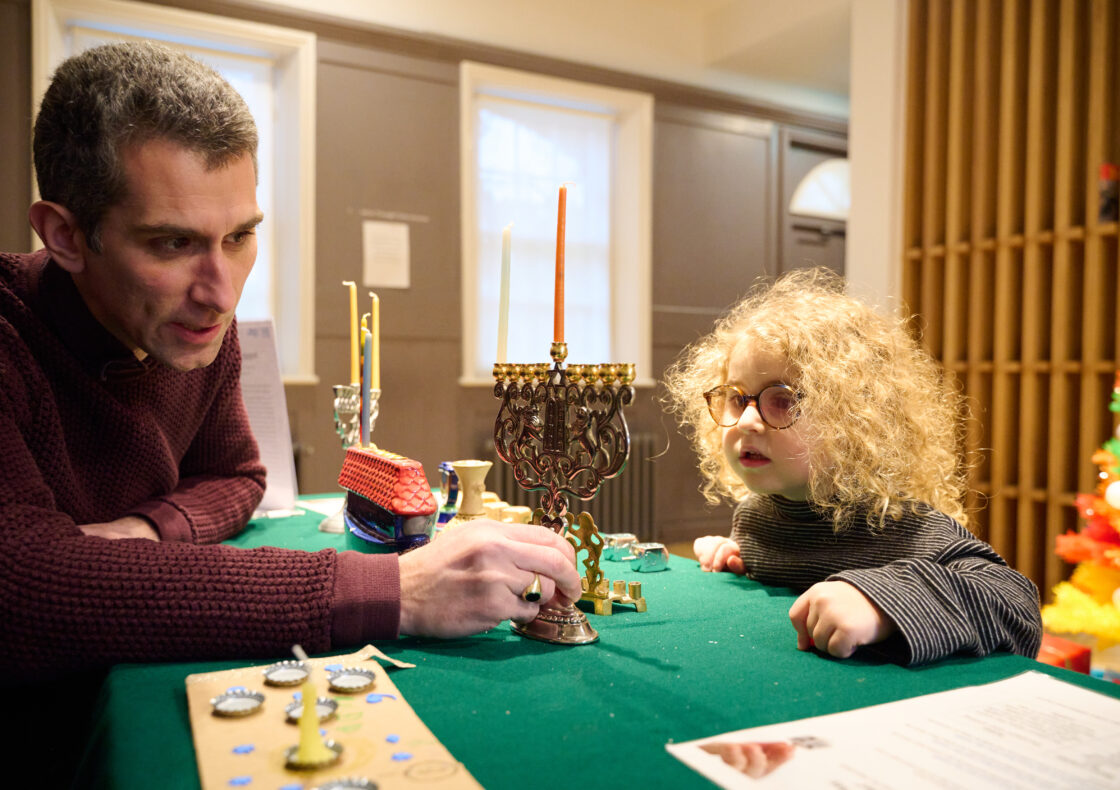 An adult speaks to a child about a multicoloured menorah on a table