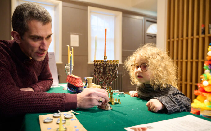 An adult speaks to a child about a multicoloured menorah on a table