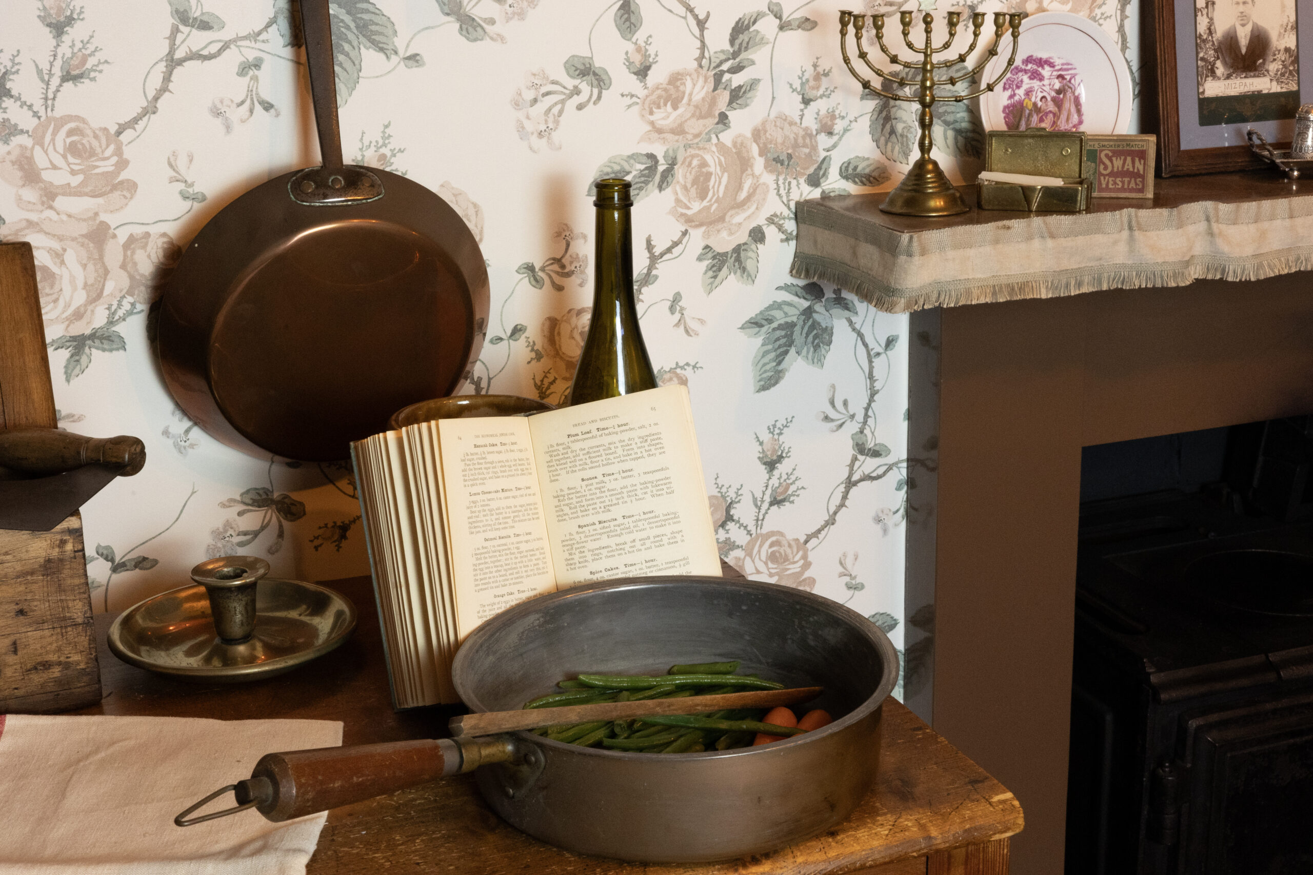 Close up view of a pot with ingredients and a recipe book. There is a menorah on the fireplace mantel.