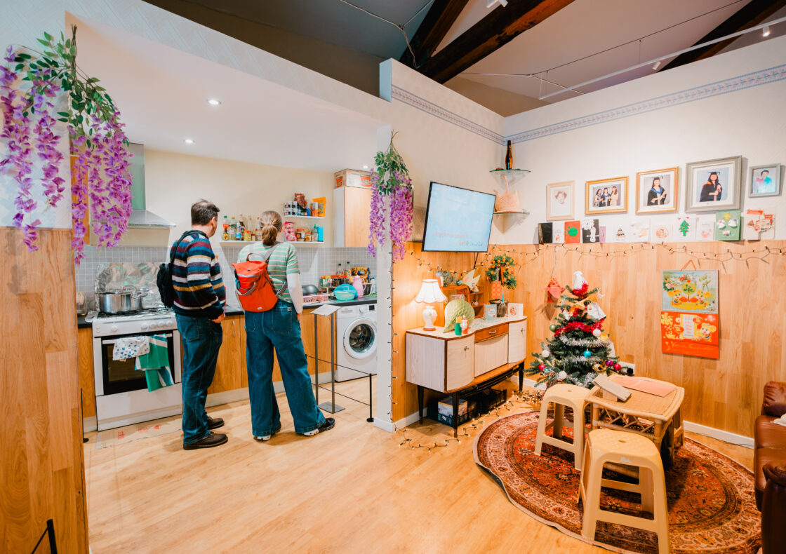 Two people standing in a room set of a living room and kitchen