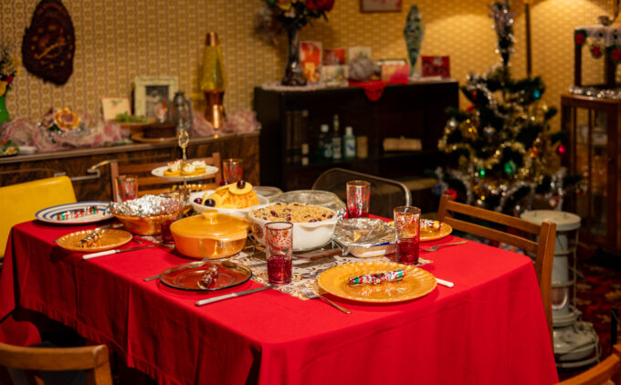 A festive dinner table set with red table cloth and warm cooked meal. There is a Christmas tree in the background.