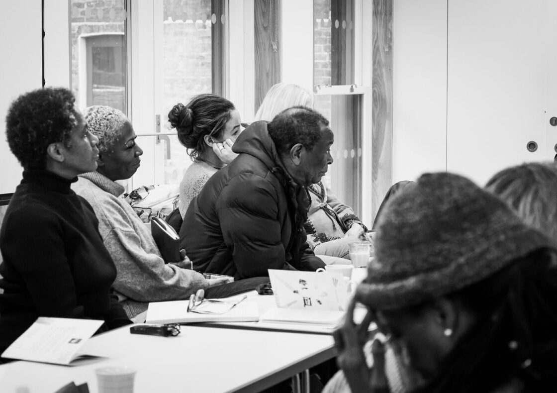 A black and white photograph of a group of people in discussion by a desk.