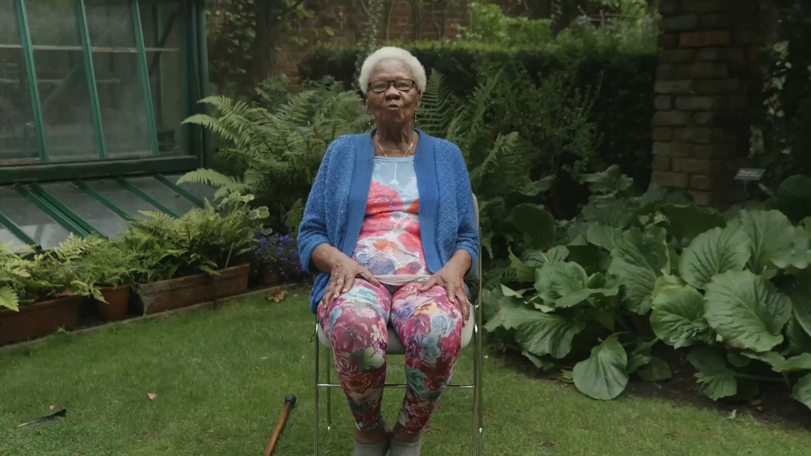 An elderly woman is sitting on a chair in a lush green garden.