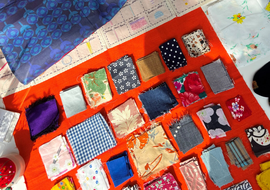 Various fabrics of different colours and patterns of cut in smaller squares place on an orange fabric. Mending items around.