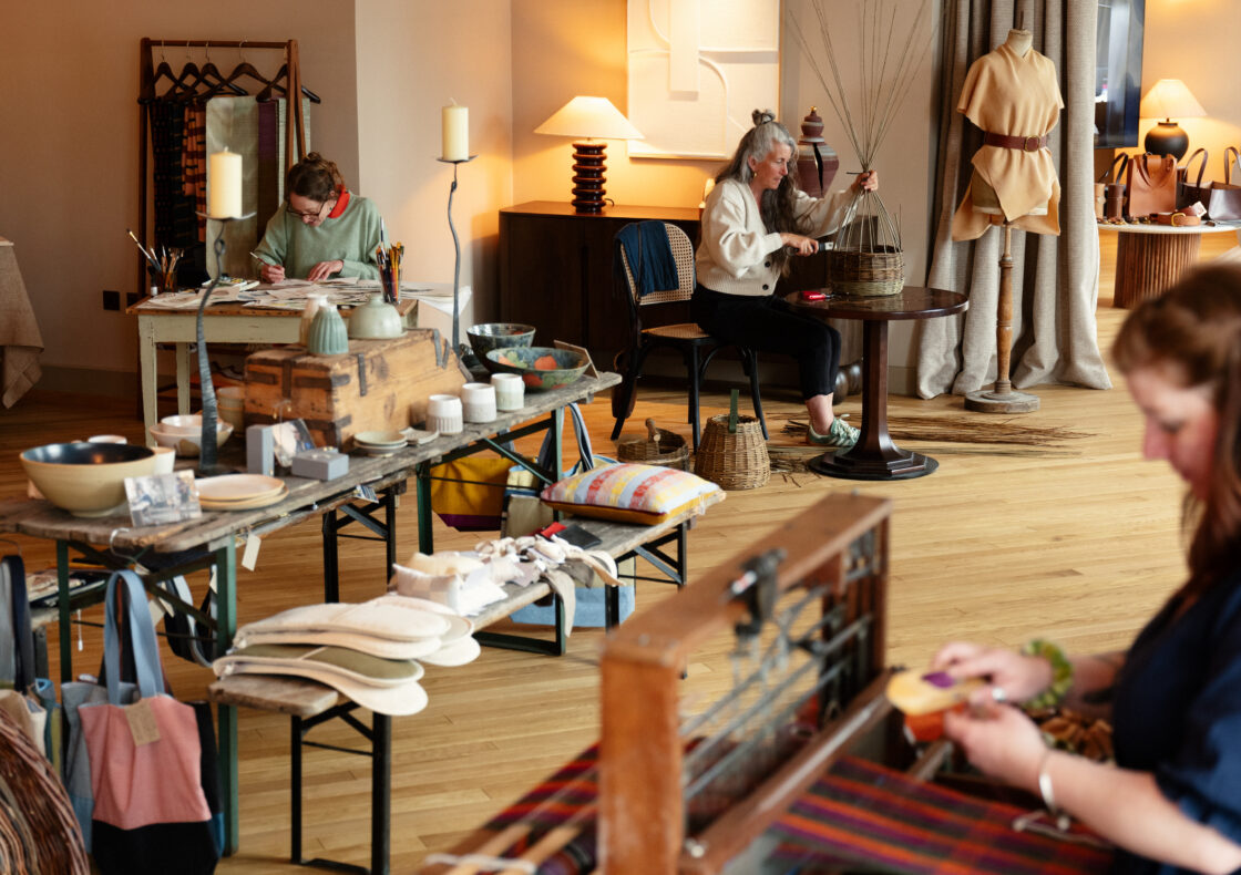 An image of a room with thee participants working on craft. One weaves textiles, one makes baskets, and one sits focussed with a paintbrush in hand. A bench in the middle has a full display of textiles, ceramics and other crafted designs.