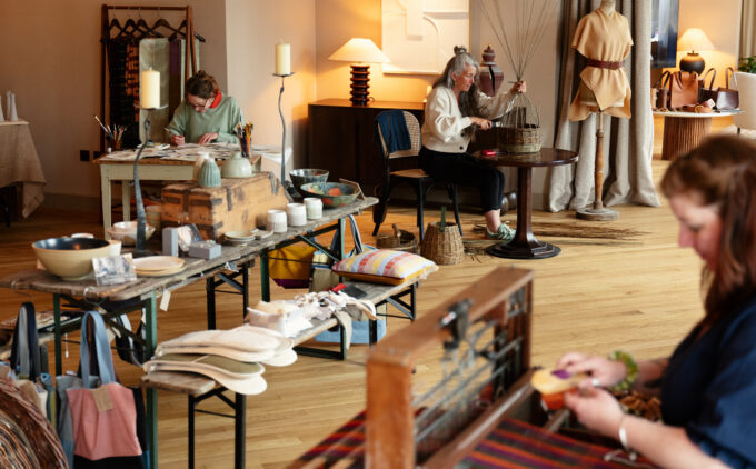 An image of a room with thee participants working on craft. One weaves textiles, one makes baskets, and one sits focussed with a paintbrush in hand. A bench in the middle has a full display of textiles, ceramics and other crafted designs.