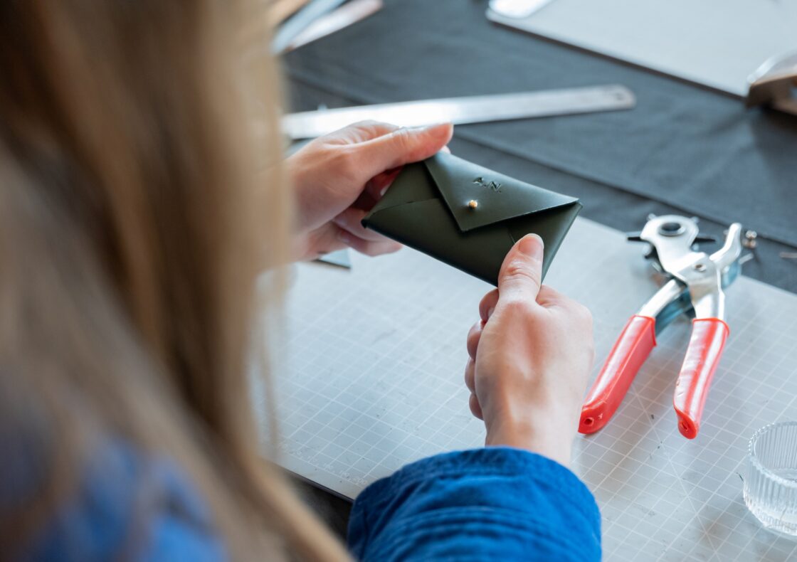 An image of hands making a leather purse with a button. Tools lie in the background.