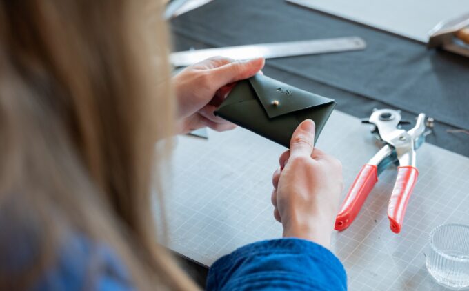 An image of hands making a leather purse with a button. Tools lie in the background.