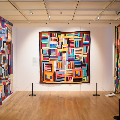 An image of a multicoloured large quilt exhibited across the wall.
