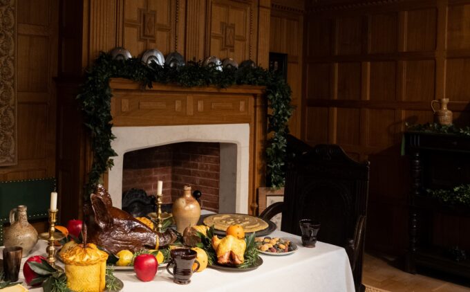 A table is laid with fruit, candles, greenery and a pigs head, in a room with wood paneling, a fireplace.